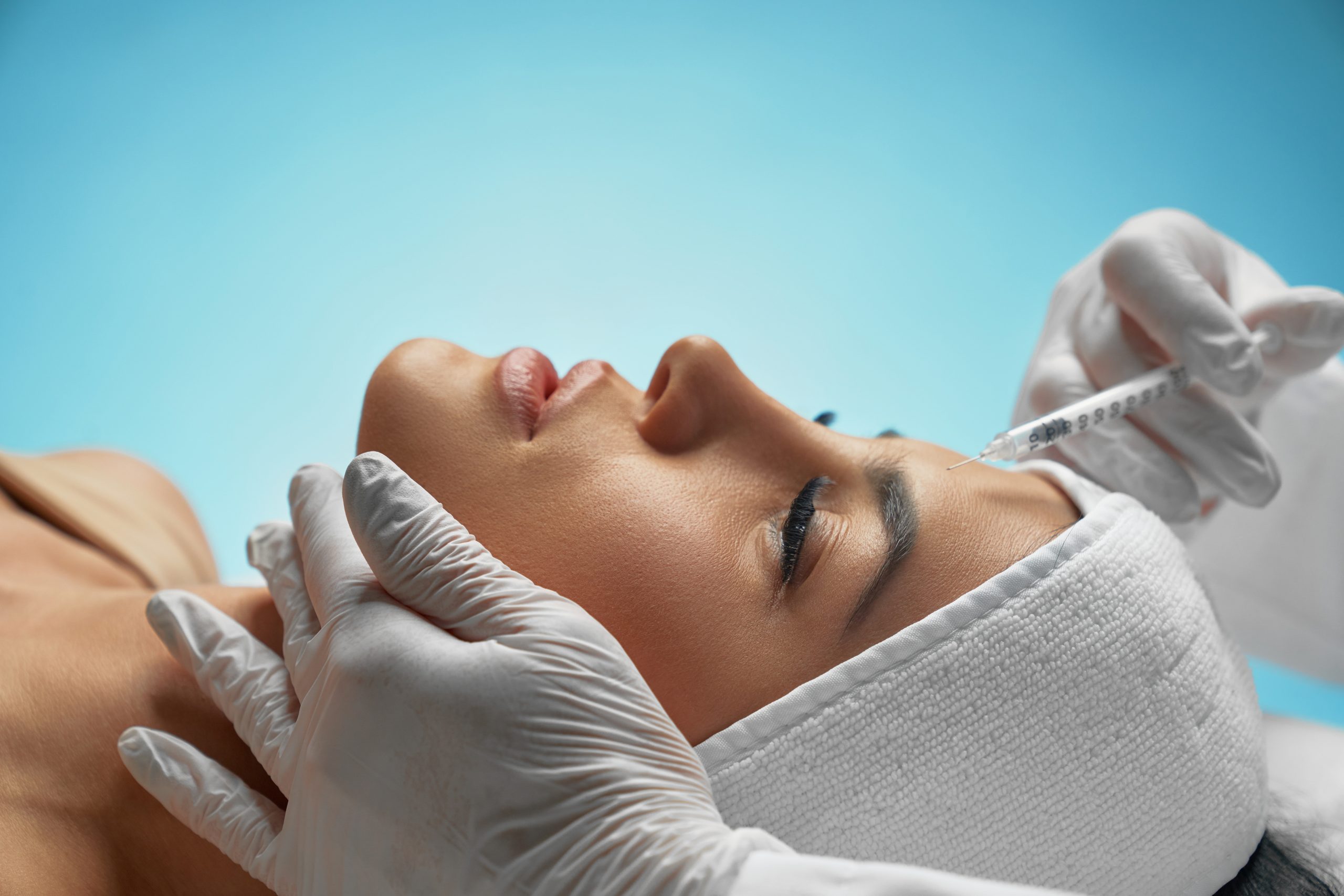 Close up of botox injection in female forehead. Side view of cosmetologist using syringe with special liquid, holding chin while patient with towel on head lying. Concept of cosmetology, beauty.